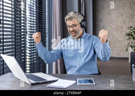 Un uomo dai capelli grigi che indossa le cuffie sta guardando le partite sportive sul posto di lavoro all'interno dell'ufficio, un gioioso capo adulto sorridente felice dei risultati. Foto Stock