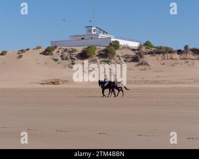 Cavalieri su una spiaggia sabbiosa con una stazione di osservazione alle spalle nella città di Essaouira, la "città ventosa", in Marocco. 20 dicembre 2023 Foto Stock