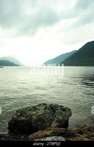 Un'enorme pietra sulla riva di un grande lago arenato tra le montagne sotto un cielo estivo nuvoloso. Lago Teletskoye, Altai, Siberia, Russia. Foto Stock