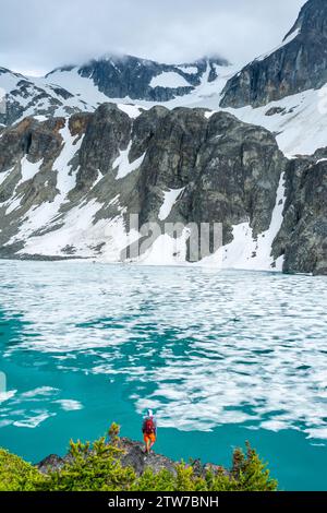 Avventuroso escursionista in rosso si affaccia sulla bellezza ghiacciata del lago Wedgemount, abbracciato da scogliere drappeggiate di neve. Foto Stock