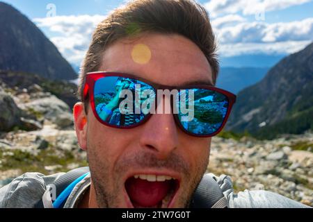 Un gioioso escursionista scatta un selfie sul Wedgemount Lake Trail con vista sulle montagne riflessa negli occhiali da sole. Foto Stock