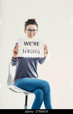 La persona allegra ha un cartello "We're Hiring", il suo sorriso luminoso che riflette la natura accogliente dell'azienda Foto Stock