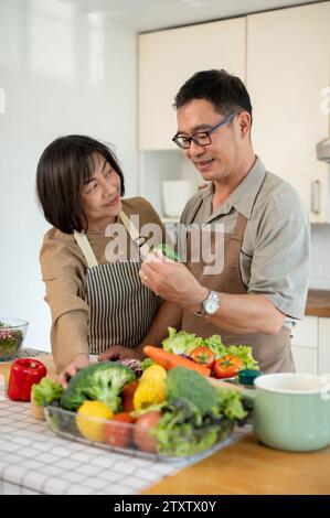 Felice moglie asiatica adulta e marito stanno cucinando la loro sana colazione in cucina insieme, parlando e preparando verdure fresche per la loro insalata Foto Stock
