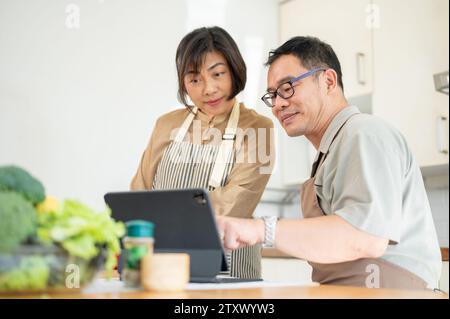 Coppie asiatiche adulte deliziose e felici stanno guardando una ricetta online su un tablet digitale e cucinando insieme in cucina. legame familiare, cucina casalinga Foto Stock