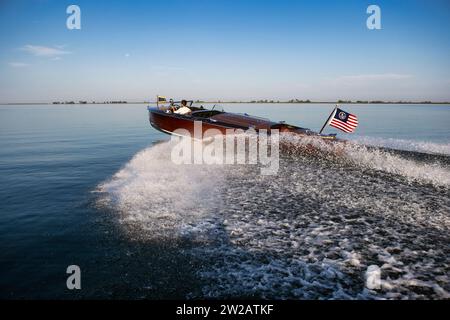 Un motoscafo in legno a tripla cabina di pilotaggio che lancia spray mentre attraversa un lago calmo. Foto Stock