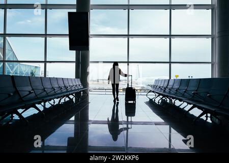 Donna con la valigia in mano, guarda fuori dalla finestra dell'aeroporto mentre aspetta il suo volo. Foto Stock