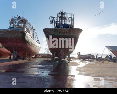 Gli uccelli volano da grandi navi e operai in un bacino di carenaggio probabilmente per manutenzione a Essaouira, la "città ventosa", in Marocco. 21 dicembre 2023 Foto Stock