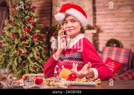 Immagine di una bella donna in abiti natalizi. Parla al telefono mentre avvolge i regali di Capodanno in una casa di campagna. Concetto di vacanza. Misto Foto Stock