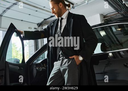 uomo elegante e attraente in un abbigliamento elegante che posa accanto alla sua auto con la mano in tasca, concetto di business Foto Stock