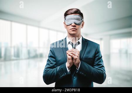 Ritratto di un uomo con un abito da lavoro. Sta pregando mentre si trova in ufficio. Ha una maschera del sonno sugli occhi. Concetto di business cieco. Supporti misti Foto Stock