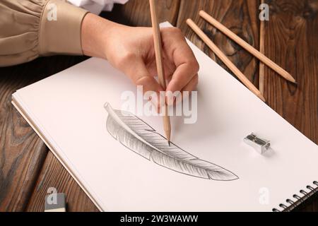 Donna che disegna piume con matita di grafite in un quaderno di bozzetti al tavolo di legno, primo piano Foto Stock