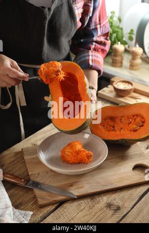 Donna che rimuove i semi dalla zucca cruda al tavolo di legno in cucina, primo piano Foto Stock