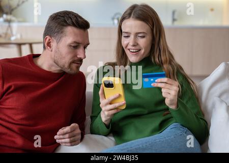 Shopping online per tutta la famiglia. Giovane donna felice con carta di credito guarda il telefono cellulare, marito sbircia Foto Stock