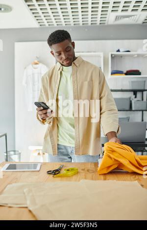 designer afro-americano con campioni di tessuto che toccano lo smartphone vicino a forbici e tablet digitale Foto Stock