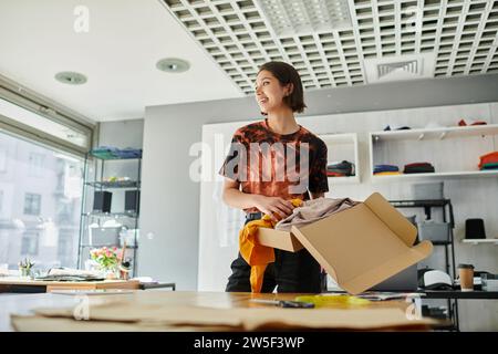 gioioso ed elegante designer asiatico che tiene una scatola di cartone con i vestiti e guarda lontano nello studio di stampa Foto Stock