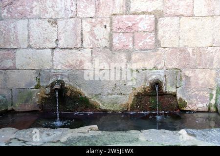 Due vecchie uscite d'acqua di una sorgente in un muro, da cui l'acqua scorre costantemente. Intorno ai rubinetti è possibile vedere le superfici ondulate sulla parete e a. Foto Stock