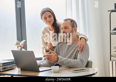 moglie premurosa con una tazza di caffè che sostiene il marito che lavora su un computer portatile in un ufficio domestico, coppia senza figli Foto Stock