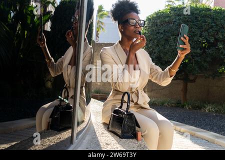 Bellissima ed elegante giovane professionista afroamericana che guarda il telefono cellulare e applica il rossetto mentre si siede all'aperto Foto Stock