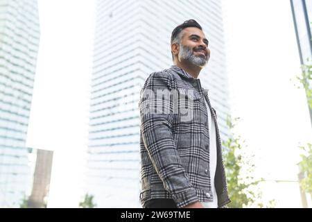 Angolo basso di uomo indiano felice che indossa abiti casual intelligenti passeggiando lungo la strada e sorridendo su un edificio moderno sullo sfondo durante il giorno Foto Stock