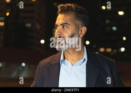 Ritratto di un uomo d'affari che indossa abiti eleganti in piedi contro le lampade luminose e che guarda fuori la sera Foto Stock