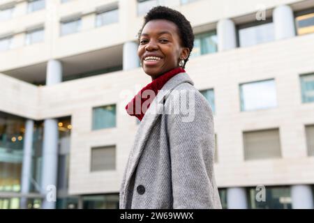 Vista laterale ad angolo basso di una giovane donna afroamericana sorridente in piedi contro edifici sfocati sullo sfondo della strada Foto Stock