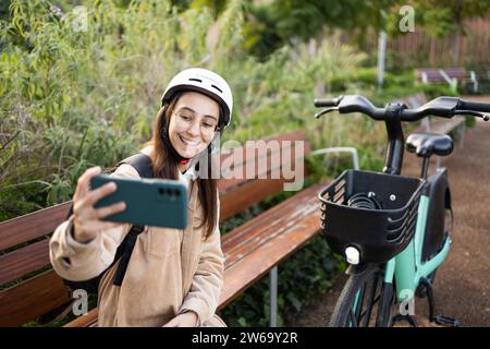 Una giovane donna sorridente scatta un selfie con il suo smartphone indossando un casco di sicurezza, in piedi accanto alla sua bicicletta in un lussureggiante parco. Foto Stock
