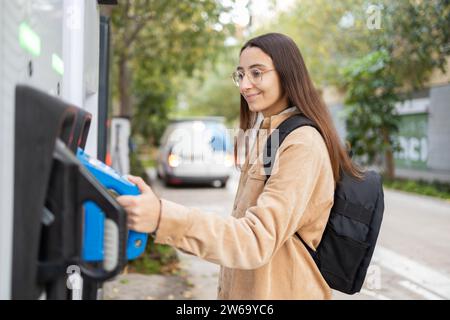 Una giovane donna sorridente indossa un abbigliamento casual usa un telefono a pagamento all'aperto in un ambiente urbano con alberi sullo sfondo. Foto Stock