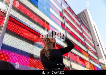 Una giovane donna elegante con occhiali da sole sorride mentre guarda in alto, in piedi di fronte a una vibrante bandiera americana esposta su uno schermo digitale a Manhattan, N. Foto Stock