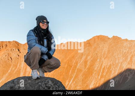 Una vera e propria turista con un sorriso toothy in occhiali da sole e abiti caldi che guardano lontano mentre si inginocchiano sulla roccia contro le scintillanti montagne gialle Foto Stock