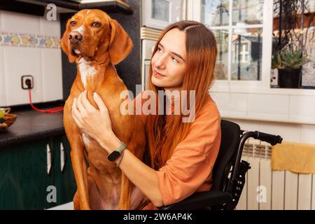 Una giovane donna in sedia a rotelle si diverte con il suo affettuoso cane in un'accogliente cucina. Foto Stock