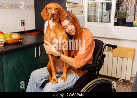 Giovane donna in sedia a rotelle che abbraccia il suo fedele cane in un'accogliente cucina casalinga. Foto Stock