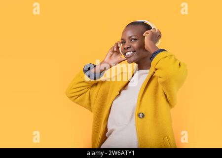 Allegra giovane donna afroamericana che guarda la fotocamera in giacca ascoltando musica attraverso cuffie wireless su sfondo giallo Foto Stock