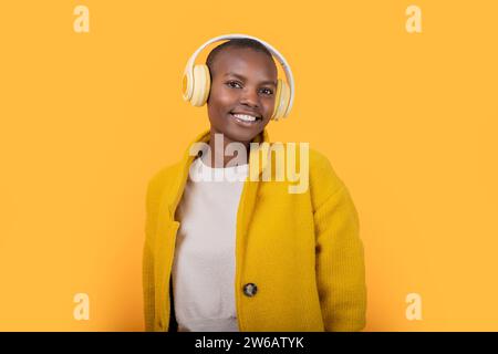 Allegra giovane donna afroamericana che guarda la fotocamera in giacca ascoltando musica attraverso cuffie wireless su sfondo giallo Foto Stock