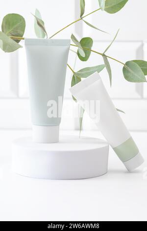 Due tubi bianchi color crema per la pelle elegantemente esposti su un piedistallo cilindrico bianco con steli in eucalipto fresco per un'estetica minimalista Foto Stock