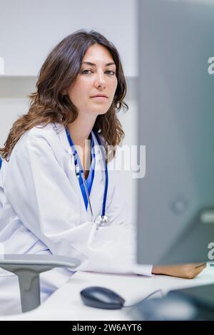 Giovane medico donna che indossa uniforme medica e stetoscopio seduto in ospedale su sfondo bianco mentre guarda la macchina fotografica Foto Stock