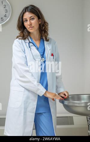 Giovane donna sicura di sé in uniforme e stetoscopio che guarda la macchina fotografica mentre si trova in una sala clinica con le mani sul lavandino contro la parete grigia con l'orologio disinserito Foto Stock