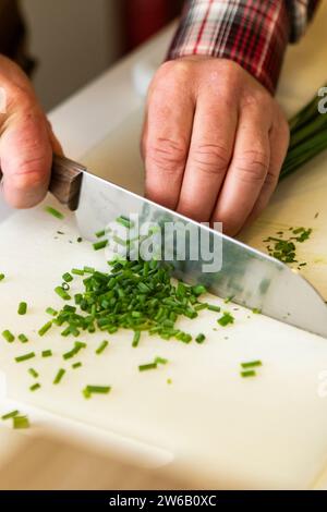 Alto angolo di ritaglio mani anonime tagliate sapientemente erba cipollina verde fresca su un tagliere bianco, con pezzi tagliati finemente sparsi acro Foto Stock