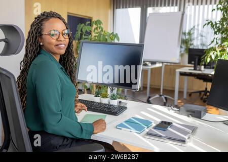 Ritratto laterale di una giovane donna d'affari afroamericana seduta alla scrivania del computer in ufficio sorridente guardando la macchina fotografica Foto Stock