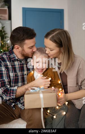 Una giovane coppia con gli occhi chiusi seduta davanti a un albero di Natale natalizio a casa, dà al bambino un bacio sulla guancia mentre guarda Foto Stock