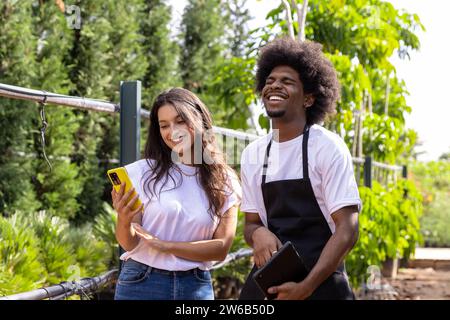 Giovane agronomo ispanico sorridente che tiene lo smartphone vicino a un collega afroamericano in piedi con un tablet digitale mentre ride in serra Foto Stock