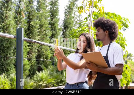 Giovane agronomo ispanico che mostra lo smartphone a un collega afroamericano in piedi con gli appunti mentre lavora insieme in serra Foto Stock