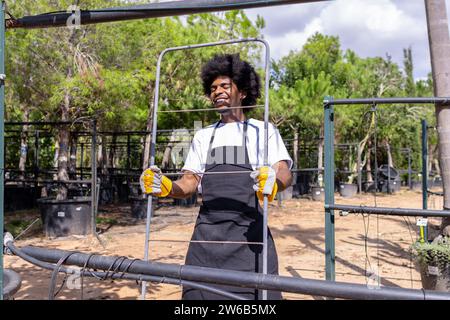 Felice giovane agricoltore afro-americano con gli occhi chiusi che trasportano una griglia metallica mentre lavorano in serra biologica contro gli alberi nelle giornate nuvolose Foto Stock