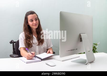 Una giovane donna professionista in sedia a rotelle siede a una scrivania con un computer moderno, prende appunti in un notebook e sorride leggermente mentre si concentra Foto Stock