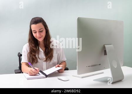 Una giovane donna professionista in sedia a rotelle siede a una scrivania con un computer moderno, prende appunti in un notebook e sorride leggermente mentre si concentra Foto Stock