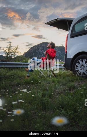 Vista laterale del turista maschile in giacca invernale che si rilassa sulla sedia vicino alla macchina su prato erboso sotto il cielo nuvoloso al crepuscolo Foto Stock