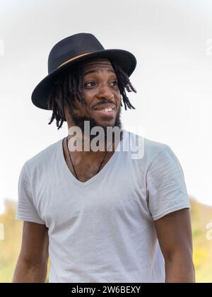 Ritratto di un giovane afro-americano barbuto con dreadlocks in cappello e maglietta bianca che guarda lontano dalla natura sfocata Foto Stock
