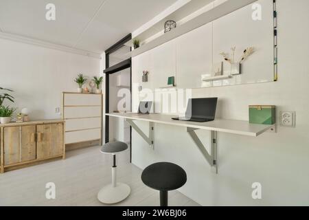 Un ufficio domestico elegante e minimalista con una scrivania bianca montata a parete, due computer portatili e un arredamento elegante che include piante e scaffali galleggianti. Foto Stock
