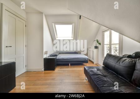 Divano in pelle nera accanto alla finestra e al letto con cuscino sotto il lucernario in una spaziosa camera da letto moderna Foto Stock