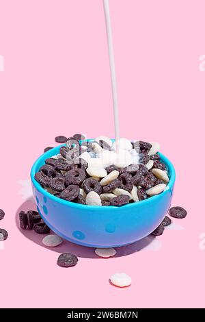 Un flusso di latte viene versato in una ciotola blu brillante ripiena di cereali al cioccolato su sfondo rosa. Foto Stock