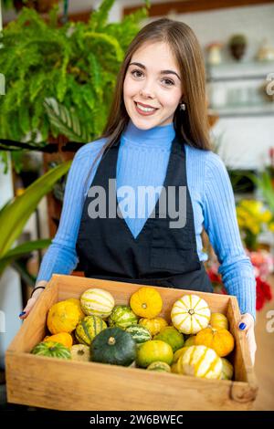 Giovane fioraio con capelli scuri che trasportano piccole zucche piene di cassa di legno mentre lavora in un fioraio Foto Stock